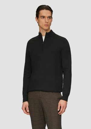 Pull-over noir avec un col zippé à moitié, fabriqué en tissu doux ; présente des manches longues et une silhouette ajustée, porté sur un pantalon écossais.