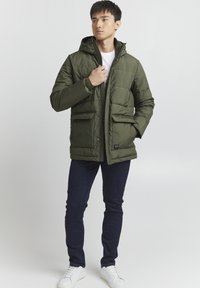 Olivenfarget polstret jakke med hette, med to frontlommer og glidelås. Brukt med mørke jeans og hvite sneakers.
