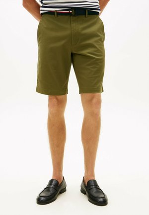 Mand iført olivengrønne shorts, sort bælte, stribet skjorte og sorte lædermokkasiner står mod hvid baggrund.