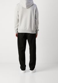 Felpa grigia con cappuccio, indossata con pantaloni neri. L'outfit presenta una vestibilità comoda e sneakers bianche. Lo sfondo è bianco semplice.