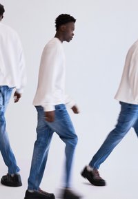 Witte langemouwen shirt, blauwe denim jeans en zwarte schoenen. Het model is in beweging, wat een vervaagd effect creëert tegen een lichte achtergrond.