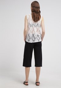 Top sin mangas de encaje blanco con patrones florales, combinado con pantalones negros anchos y recortados; diseño minimalista, textura suave y sandalias sencillas.