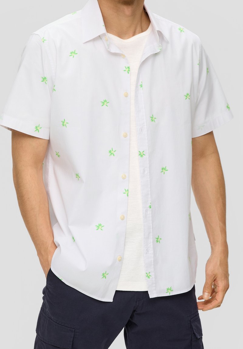 Homme portant une chemise blanche à manches courtes avec un petit imprimé floral vert sur un t-shirt blanc et un short cargo foncé.