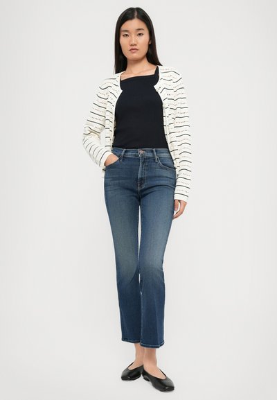 Wit gestreepte cardigan over een zwarte top, gecombineerd met donkerblauwe jeans en zwarte flats. Beschikt over een getailleerd ontwerp met recht gesneden pijpen.