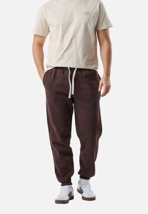 Homme portant une chemise beige à manches courtes avec un petit imprimé, un pantalon de jogging marron foncé avec cordon de serrage, et des baskets blanches à rayures noires.