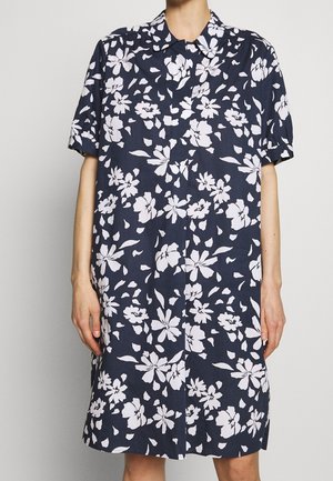 Osoba ubrana w granatową, krótką sukienkę z krótkim rękawem, sięgającą do kolan, z białym kwiatowym wzorem i zaostrzonym kołnierzykiem.