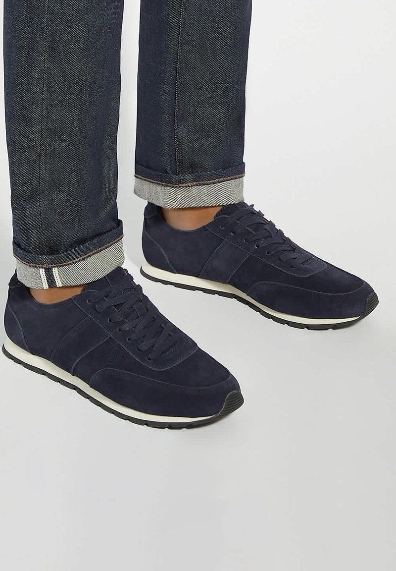 Navy suede athletic shoes with a white midsole and black outsole. Features include rounded toe, lace-up design, and contrasting fabric accents.