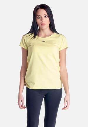 Giovane donna con lunghi capelli scuri, indossa una maglietta giallo chiaro a maniche corte e leggings neri, in piedi davanti a uno sfondo semplice.