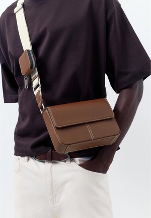 Homme portant une chemise marron foncé et un pantalon blanc, portant un sac bandoulière en cuir marron avec une sangle blanche et une petite pochette attachée.
