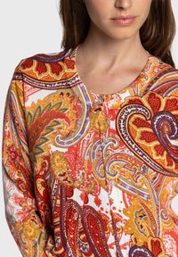 Blouse colorée à motif cachemire en rouge, orange et jaune, avec un col rond et des boutons à l'avant. Texture de tissu douce.