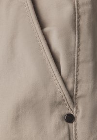 Beige bomullstyg med en texturerad yta, som har en sned söm och en svart tryckknapp för stängning.