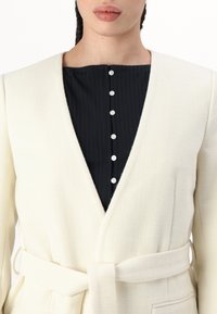 Veste enveloppante de couleur crème, texturée, avec une ceinture, superposée sur un haut noir à côtes avec un col rond et six boutons blancs.