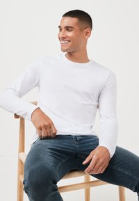 Hvid langærmet skjorte lavet af bomuld, rund halsudskæring, parret med mørkeblå jeans. Casual pasform, siddende på en træstol.