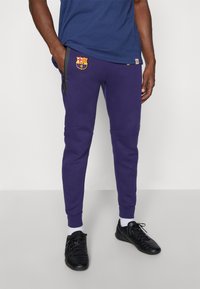 Paarse trainingsbroek met een Barcelona-logo, voorzien van een trekkoord, zijzakken en geribbelde enkels. Draagt zwarte sportschoenen.