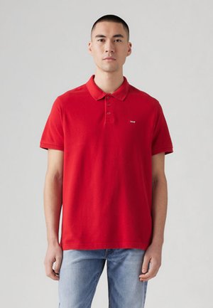 Hombre con una camiseta polo roja lisa de manga corta y vaqueros azul claro, de pie frente a un fondo liso de color gris claro.