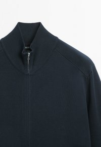 Granatowy dzianinowy sweter z wysokim prążkowanym kołnierzem i krótkim srebrnym zamkiem z przodu, pośrodku.