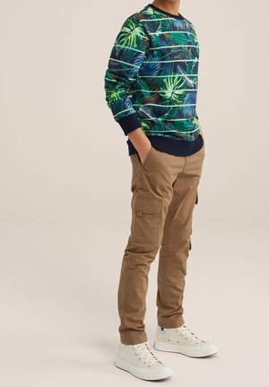 Garçon portant une chemise à manches longues à motifs de feuilles vertes et bleues, un pantalon cargo beige et des baskets blanches montantes, les mains dans les poches.