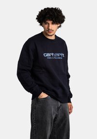 Sudadera azul marino con puños y dobladillo acanalados, que presenta un logotipo bordado en azul y blanco "carhartt WORK IN PROGRESS" en la parte frontal.