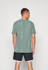 Szürke-zöld, rövidujjú sportpóló, texturált kötésű mintával és függőleges "UNDER ARMOUR" logóval a hátsó részen. Fekete sorttal viselve.
