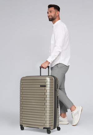 Valise rigide en argent avec des rainures verticales, une poignée rétractable et quatre roues. Texture lisse et logo visible à l'avant.