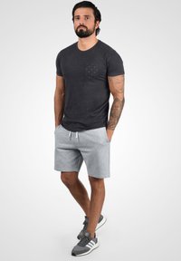 T-shirt à manches courtes gris foncé avec une poche à motifs, associé à un short gris clair et des chaussures de sport grises avec des semelles blanches.