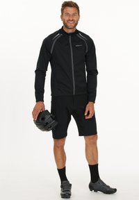Svart cykeljacka med reflexdetaljer, ihop med svarta shorts. Modellen håller en svart hjälm, har på sig svarta strumpor och cykelskor.