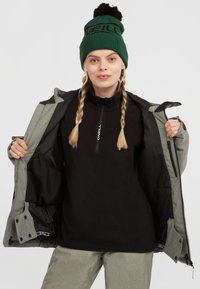 Zwarte fleecetrekker met een ritskraag, gedragen onder een grijze jas. Het model draagt een groene gebreide muts met een bruine pompon en print.