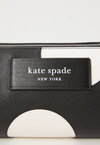 Svart skinnlommebok med en hvit sirkulær detalj, preget logo med teksten "kate spade NEW YORK," og en sølvfarget glidelåsdetalj.