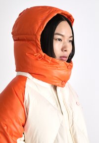 Giacca imbottita con cappuccio arancione e corpo crema. Presenta una chiusura con zip, dettagli con bottoni e una texture trapuntata.