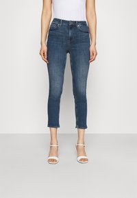 Jeans de denim en azul oscuro, de tiro medio, ajustados y con una longitud corta. Presentan bolsillos delanteros y costuras blancas. Se llevan con sandalias blancas de tiras.