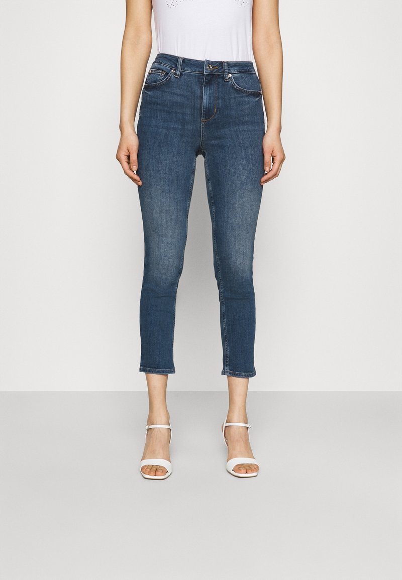 Jeans de denim en azul oscuro, de tiro medio, ajustados y con una longitud corta. Presentan bolsillos delanteros y costuras blancas. Se llevan con sandalias blancas de tiras.