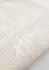 Kudumine kreemjas beanies, millel on tekstuurne muster, esiküljel on volditud servas õmmeldud logo sobivas värvitoonis.