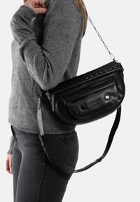 Zwarte leren schoudertas met een ritssluiting, studs als accent en een getextureerde voorkant, gedragen over een grijze trui en zwarte jeans.