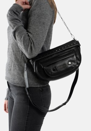 Sac en cuir noir porté en bandoulière, avec une fermeture éclair, des accents cloutés et une poche avant texturée, porté sur un pull gris et un jean noir.
