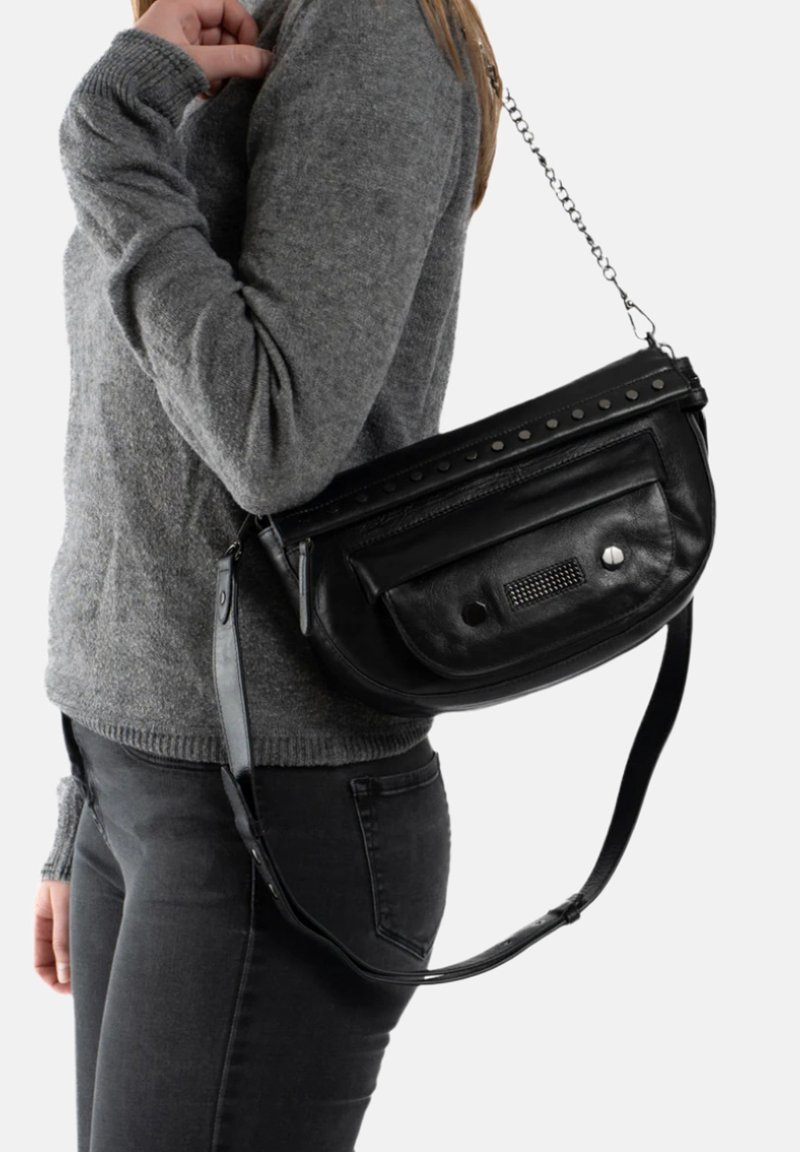 Zwarte leren schoudertas met een ritssluiting, studs als accent en een getextureerde voorkant, gedragen over een grijze trui en zwarte jeans.