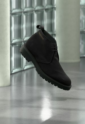 Schwarzer Wildleder-Stiefelette mit dicker profillierter Sohle und Zuglasche, seitlich mit goldenem Valentino-Schriftzug, schwebend über poliertem Betonboden.