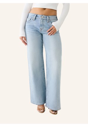 Personne portant un jean bleu clair taille haute à jambes larges, un crop top blanc à manches longues et des sandales à bout ouvert, debout devant un fond uni.