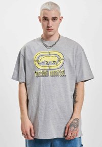 Lichtgrijze t-shirt met korte mouwen, voorzien van een geel neushoornlogo en zwarte tekst. Gemaakt van katoen, met een relaxte fit en zachte textuur.