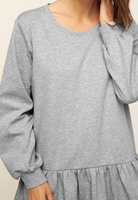 Femme portant un haut à manches longues gris clair avec des détails froncés à la taille, main touchant son cou, fond neutre.