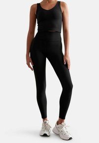 Top corto senza maniche nero con scollatura a V e leggings neri a vita alta. Modella con sneaker bianche. Tessuto morbido, design aderente.