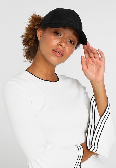 Casquette de baseball noire avec un logo en relief, portée avec un haut blanc comportant des accents rayés noirs sur les manches. Cheveux bouclés partiellement visibles.