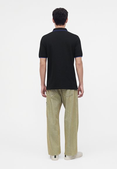Homme debout de dos, portant un polo noir avec une bordure bleue, un pantalon beige ample et des baskets blanches sur un fond blanc uni.