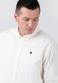 Weißes Cordhemd mit Knopfleiste und kleinem schwarzem Logo auf der linken Brust, mit klassischem Kragen und strukturierter Stoffoberfläche.