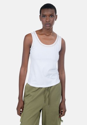Donna che indossa una canottiera bianca con borchie dorate intorno al collo e pantaloni cargo verde oliva, in piedi davanti a uno sfondo semplice.
