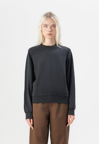 Sweatshirt negru din bumbac cu mâneci raglan, cu mansete și tiv din material ribat. Stilizat cu pantaloni largi maro. Textură netedă, design minimalist.