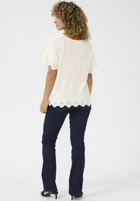 Top bianco lavorato a maglia con maniche corte e orlo in pizzo a scallop, abbinato a jeans svasati blu scuro. Capelli ricci visibili da dietro.