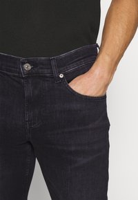 Mies, joka pukeutuu tummiin farkkuihin ja jonka käsi on osittain oikeassa etutaskussa, on pukeutunut mustaan tekstiiliin. 