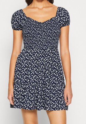 Robe courte bleu marine avec motif floral blanc, corsage smocké, manches bouffantes et jupe à volants portée par une femme.