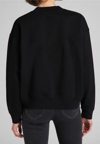 Persona con una sudadera negra que tiene una costura de yugo curvada en la parte superior de la espalda y jeans gris oscuro, de pie contra un fondo gris.