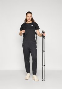 Crna sportska majica i trenirke, oba s logotipom. Drži crne planinarske štapove, nosi svijetle planinarske cipele.